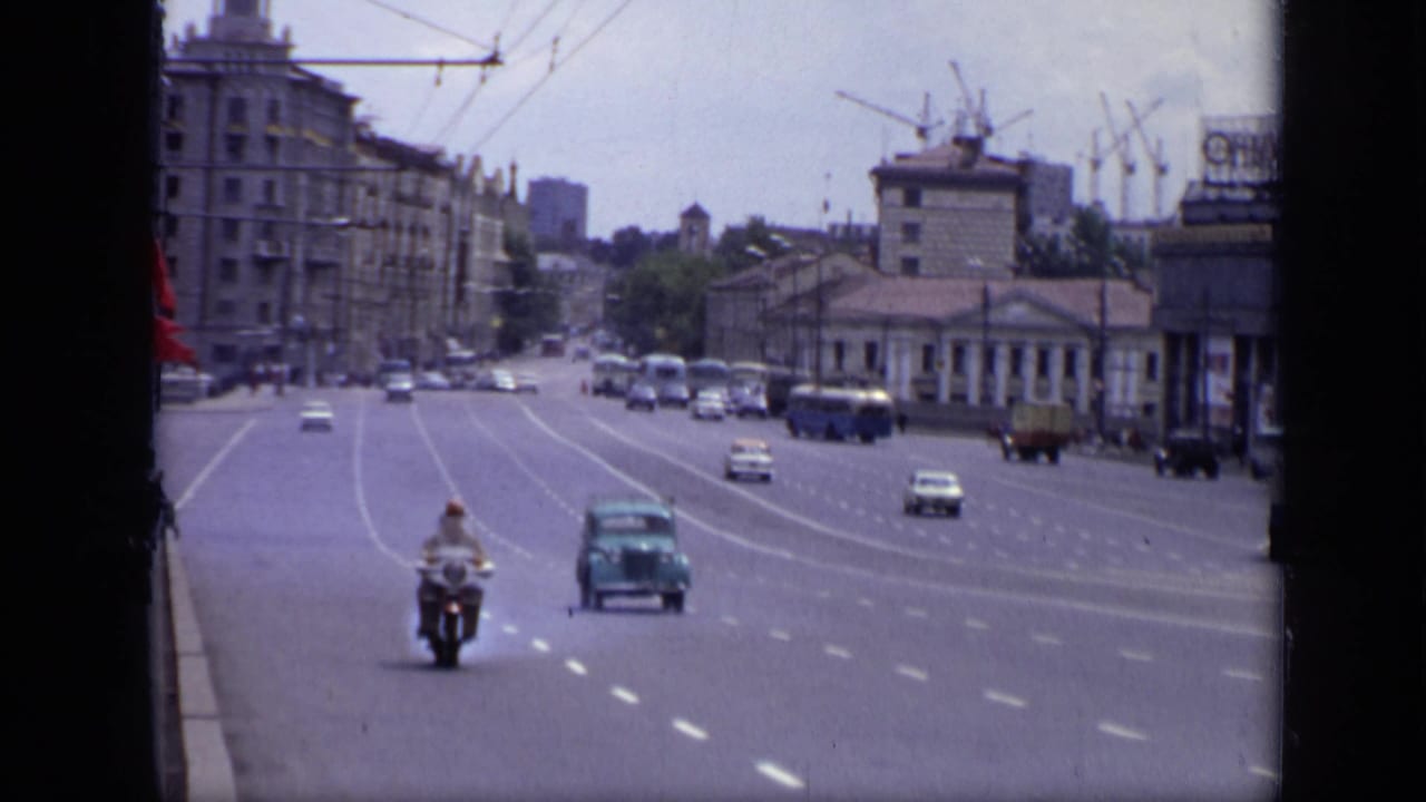 Wide Moscow road with motorcycle, cars, buildings, trolley wires, and distant cranes.