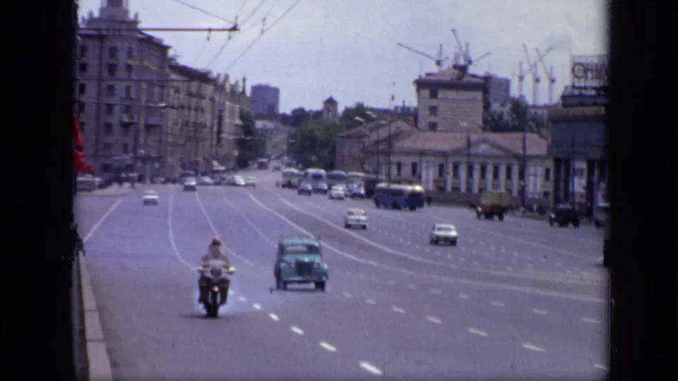 1971 Moscow street view with a motorcyclist approaching, cars on a broad avenue, overhead wires, red flags, buildings, and cranes.