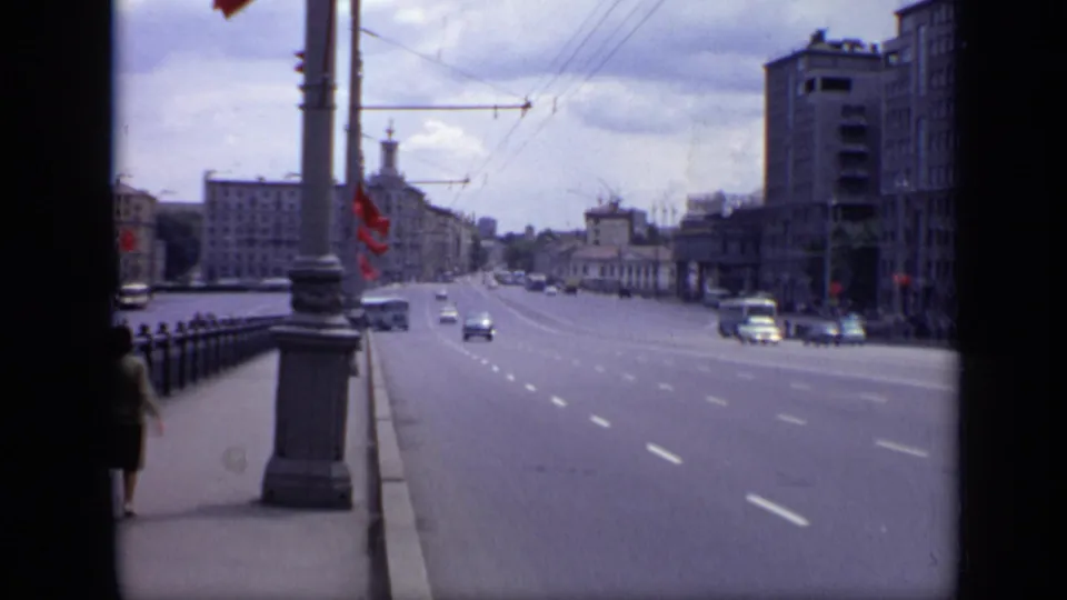 1971 Moscow street scene with cars on a broad road, a pedestrian partly visible on the sidewalk, red flags, wires, and large buildings.