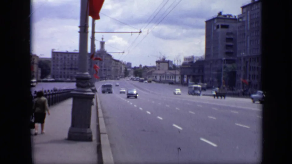 1971 Moscow road traffic with several cars, small pedestrian figures, red flags on poles, overhead wires, and tall urban buildings.