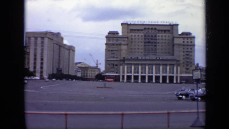 1971 Moscow street frame showing a prominent columned building, adjacent blocky structure, Cyrillic rooftop signage, sparse cars, and foreground railing.