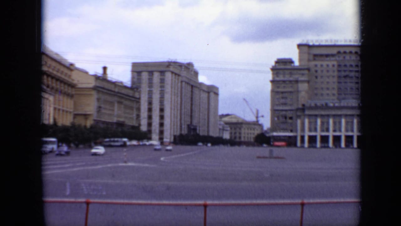 Wide city square with Soviet-era buildings, cars, buses, and a red roadside railing.