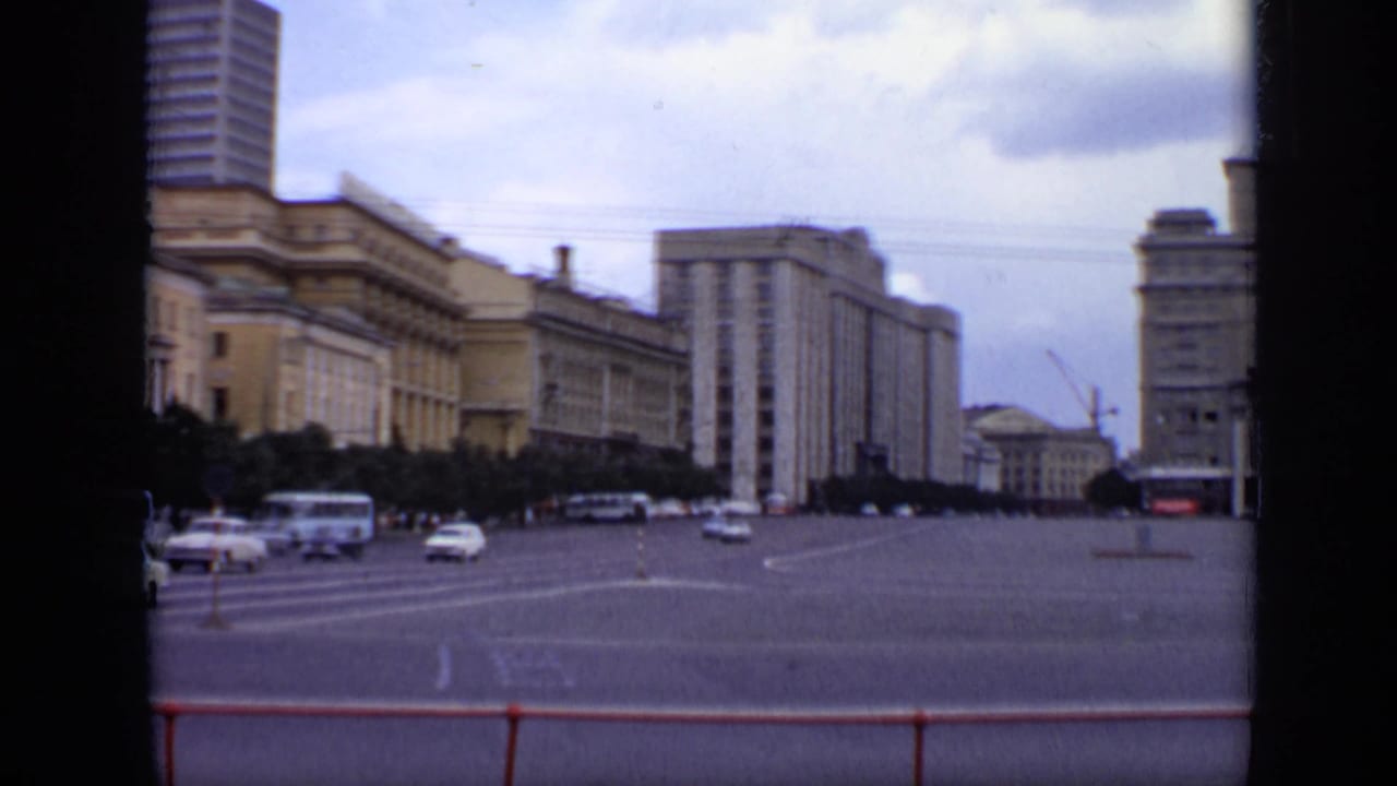 Broad road and city buildings with cars, buses, and overhead wires under cloudy sky.