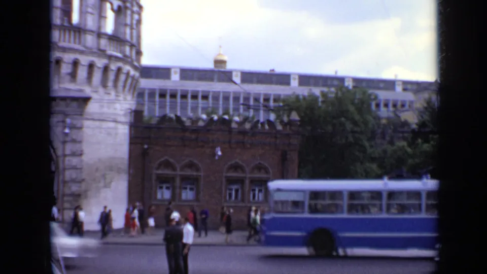 1971 Moscow street view with pedestrians near a stone tower, a blue bus crossing the frame, and civic buildings behind it.