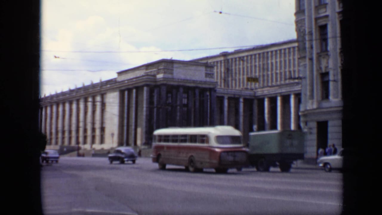A bus, cars, and a box truck move past a large columned urban building.
