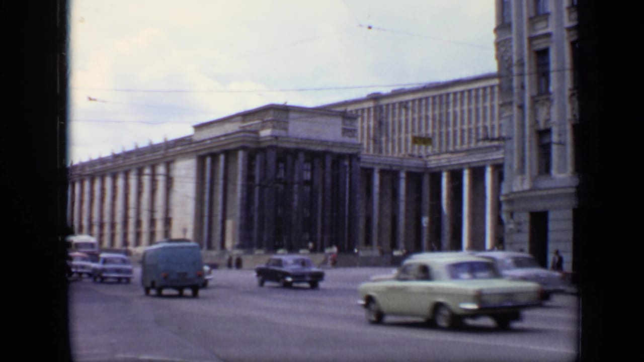 Cars and a van move along a wide street in front of a large stone building.