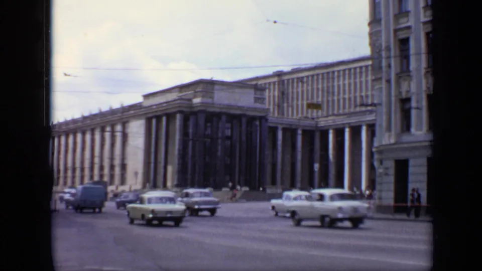 1971 Moscow traffic scene with pale sedans and a small van moving below a monumental colonnaded facade.