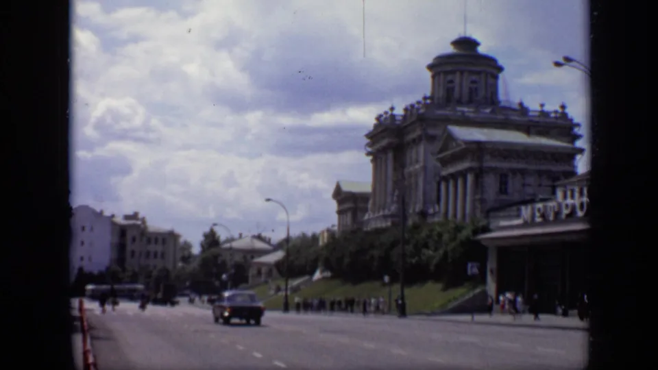 1971 Moscow street scene with a dark car on the road, pedestrians near the Metro entrance, and the domed building in the background.