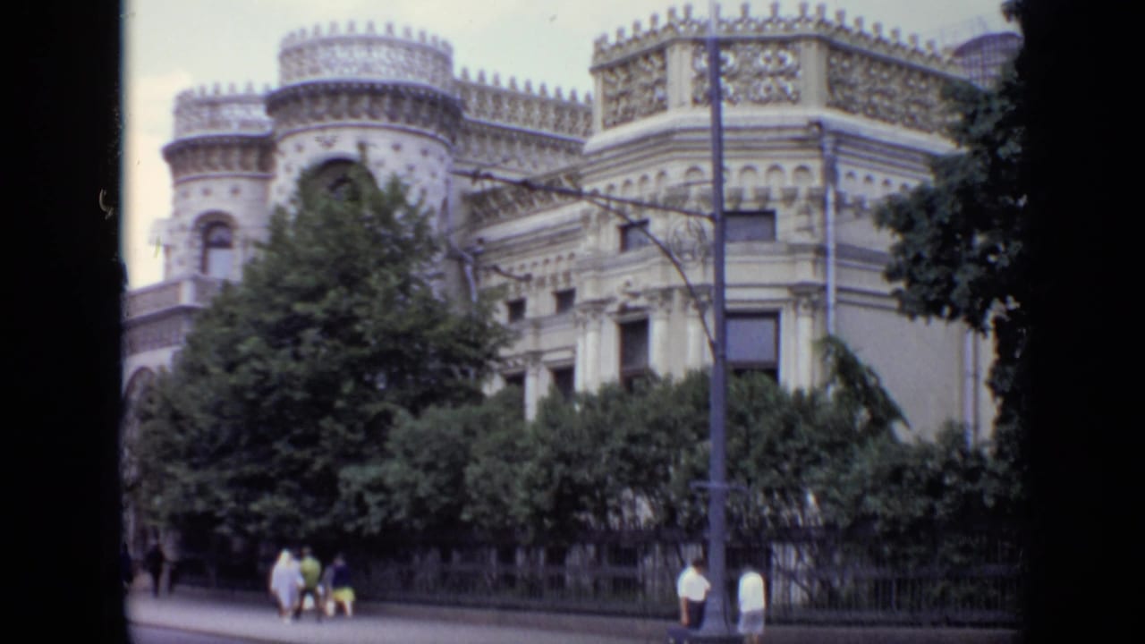 1971 Moscow street view with a decorative historic facade, trees, sidewalk pedestrians, and a street lamp in soft 8mm color.