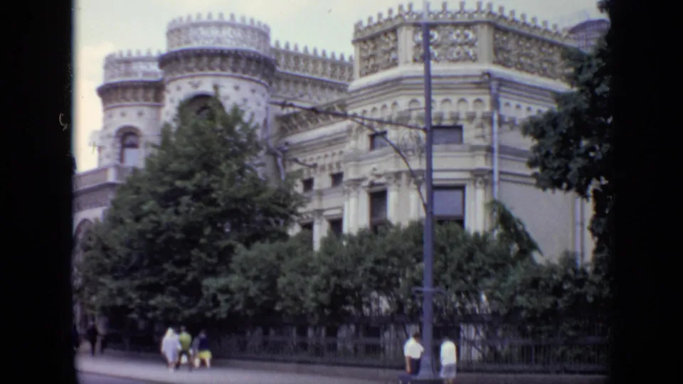 1971 Moscow street view with a decorative historic facade, trees, sidewalk pedestrians, and a street lamp in soft 8mm color.