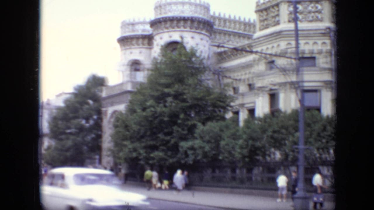A light-colored period car moves along a Moscow boulevard in front of trees, pedestrians, and ornate historic architecture.