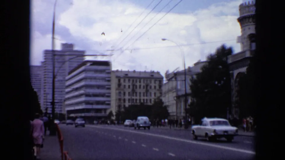 Wide Moscow street scene showing moving cars, pedestrians, overhead wires, street lamps, and mid-century buildings under a cloudy sky.