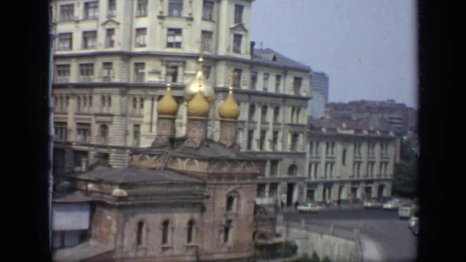 1971 Moscow home-movie frame showing church domes, dense city buildings, a curving street, small cars, and distant pedestrians.