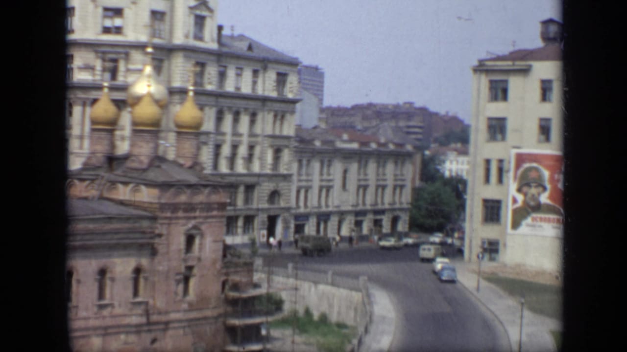1971 Moscow street scene showing golden onion domes, Soviet-era buildings, light traffic, pedestrians, and a large soldier poster on a facade.