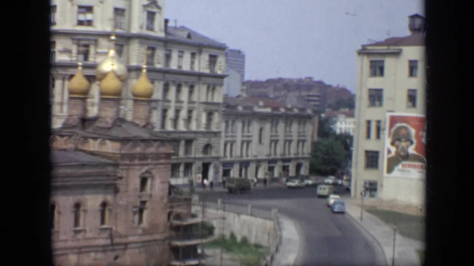 1971 Moscow street scene showing golden onion domes, Soviet-era buildings, light traffic, pedestrians, and a large soldier poster on a facade.