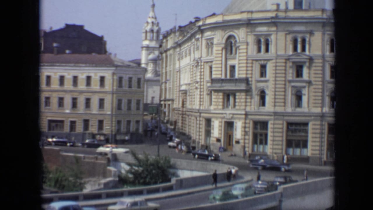1971 Moscow street view with ornate facades, period cars, pedestrians, a curved road barrier, and a church spire in the background.