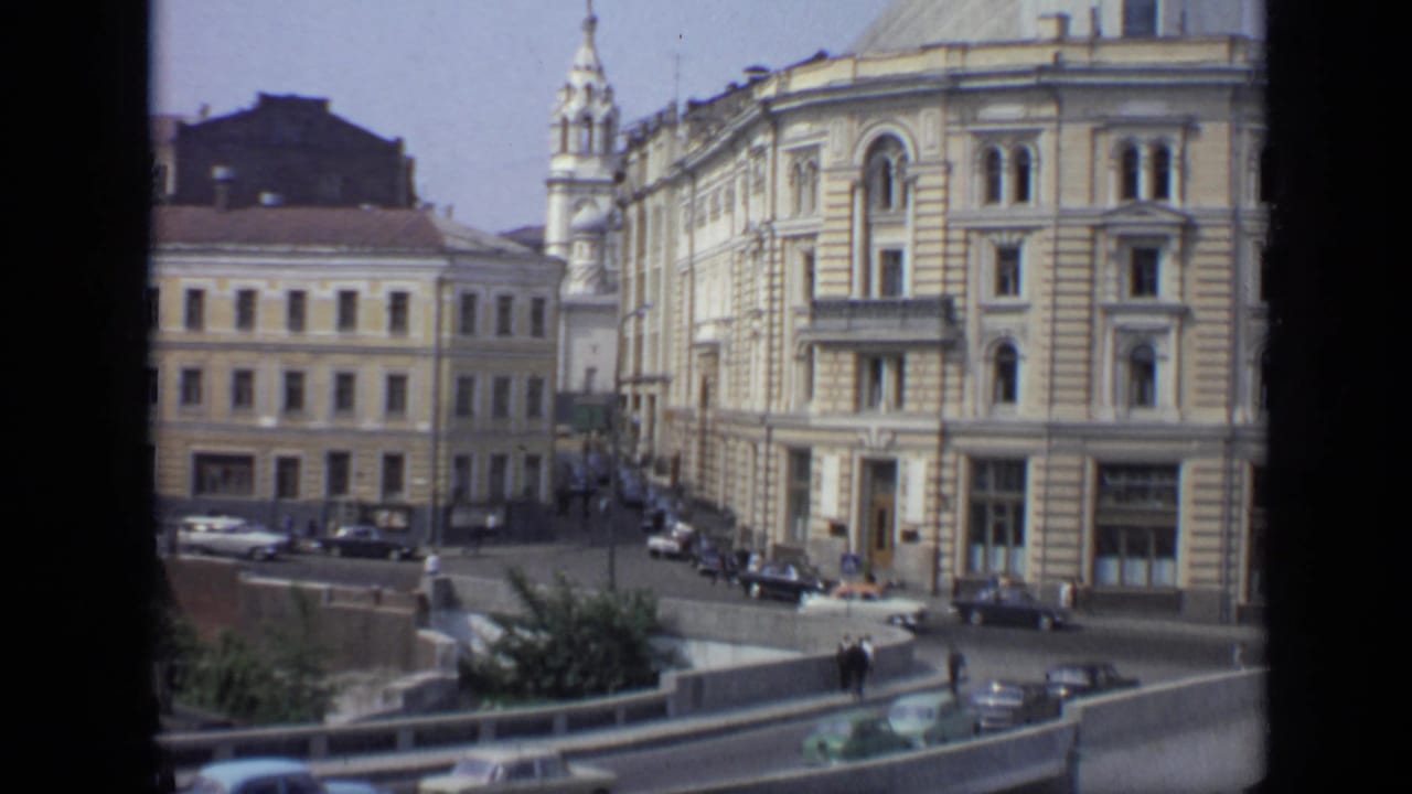 1971 Moscow view of a formal urban block, with parked and moving cars, pedestrians, sidewalks, and a church spire behind the buildings.