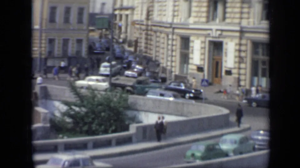 Tighter 1971 Moscow street view with period cars, pedestrians, ornate building entrances, a curved road barrier, and greenery in the foreground.