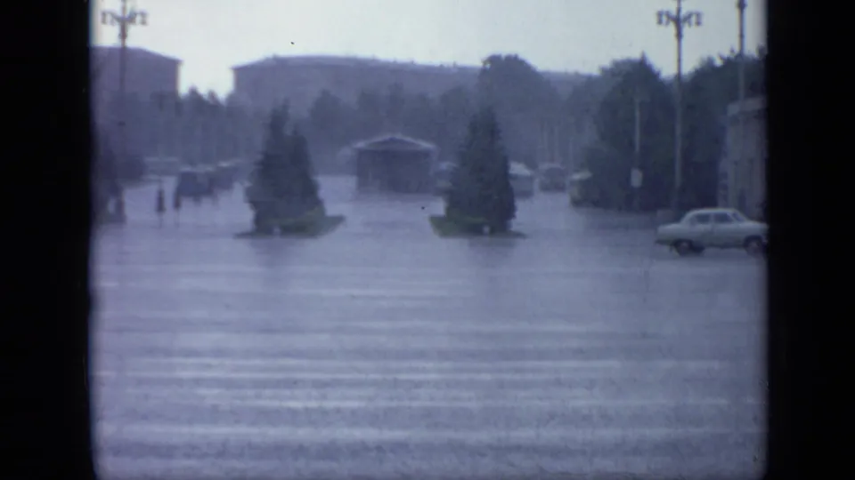Rainstorm view in Moscow showing a flooded roadway or intersection, trees, utility poles, distant buildings, and vintage cars.