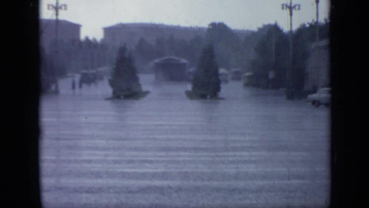 Flooded city street with crosswalk, small planted islands, streetlamps, and distant cars in heavy rain.