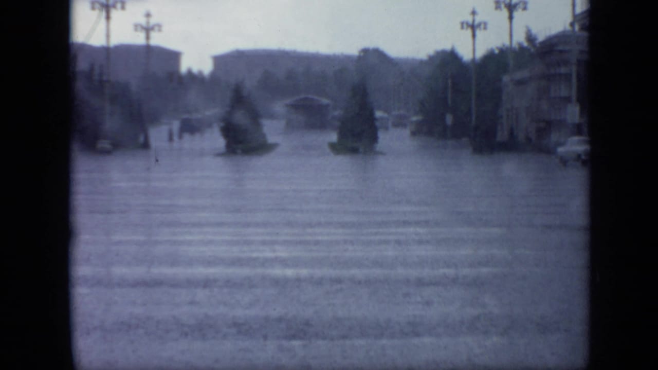 Moscow roadway in 1971 with rainwater covering the street, small tree islands, faint pedestrians, streetlamps, and a car at right.