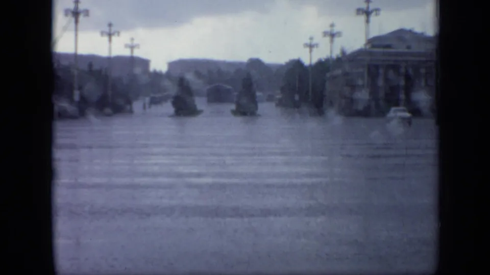 Moscow street in heavy rain, with blue-gray 8mm tones, flooded pavement, crosswalk lines, planted islands, streetlamps, and a car at right.