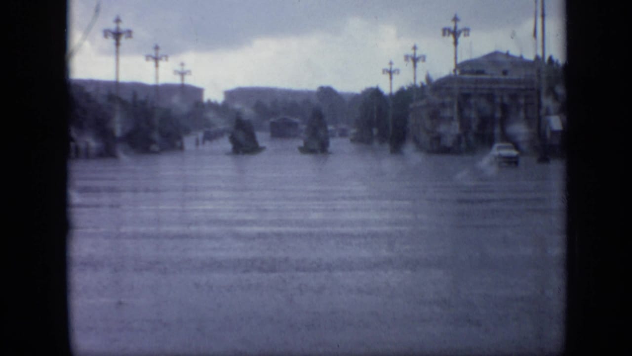 Flooded broad street with lampposts, trees, distant buildings, and a small car at right.