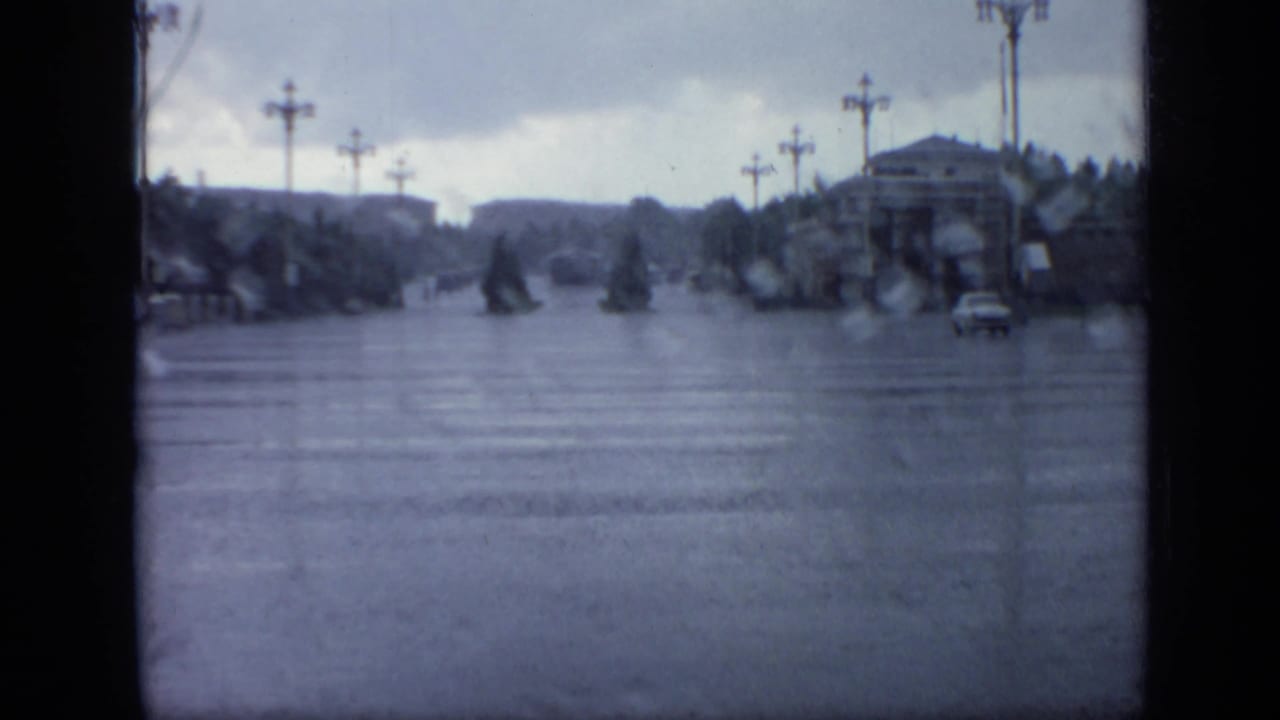 Wide flooded Moscow roadway in 1971, seen through rain-softened 8mm footage with lampposts, trees, distant buildings, and a car at right.