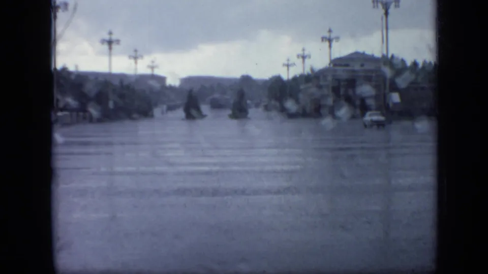 Wide flooded Moscow roadway in 1971, seen through rain-softened 8mm footage with lampposts, trees, distant buildings, and a car at right.