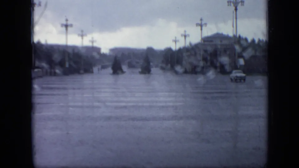 Blue-gray 8mm frame of a flooded Moscow street in 1971, with formal lampposts, trees, distant architecture, and a small car on the right.