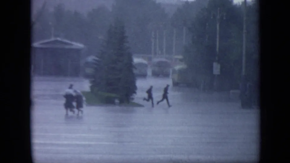 In 1971 Moscow, several distant pedestrians run through floodwater near trees and lampposts, with a small shelter structure and cars beyond.
