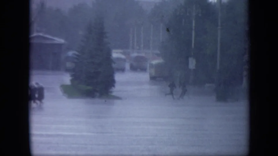 1971 Moscow home-movie frame showing pedestrians crossing a wet open street area during heavy rain near trees and distant structures.