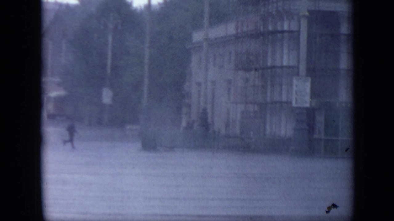 1971 Moscow frame with pedestrians crossing wet pavement in heavy rain beside a large public-looking building facade.