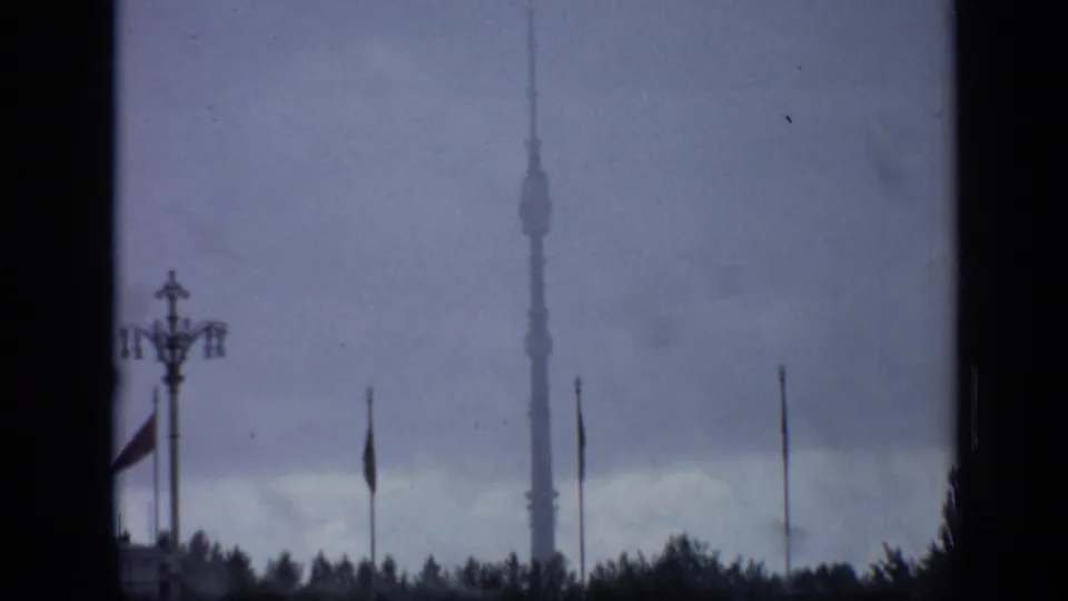 1971 Moscow home-movie frame showing a distant tower-like structure beyond trees, flags or poles, and a dark rainy sky.