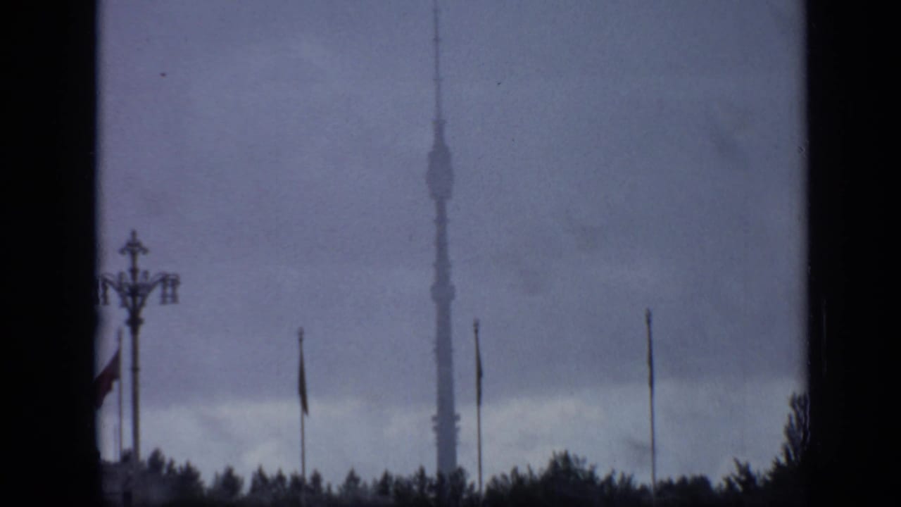 Ostankino Tower behind flagpoles, treetops, and a decorative streetlamp under a cloudy sky.