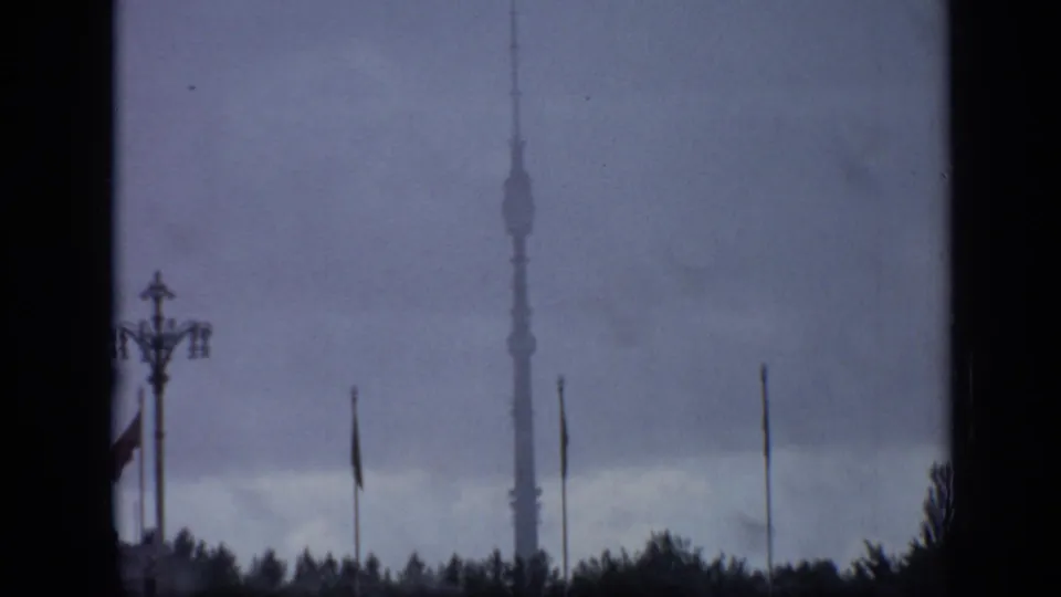 1971 Moscow view of Ostankino Tower framed by dark window edges, with flagpoles, treetops, a streetlamp, and overcast sky.