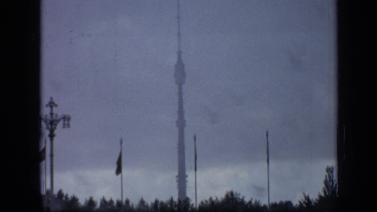 Moscow landmark view showing Ostankino Tower through dark framing, with several flagpoles, a lamppost, trees, and heavy cloud cover.