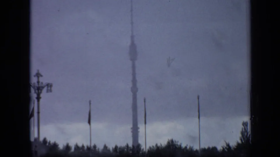 Soft 8mm Moscow view of Ostankino Tower and flagpoles beyond trees, partly obscured by dark window or structural framing.