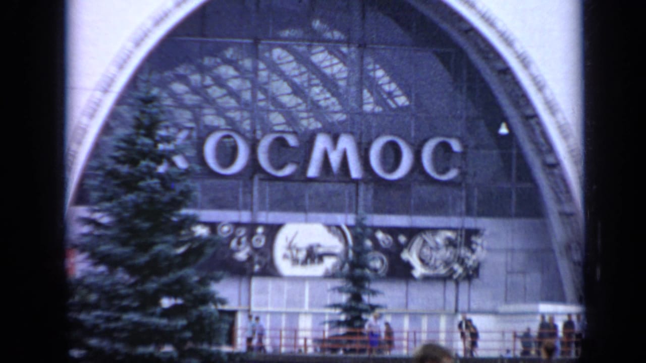 Kosmos pavilion facade with large Cyrillic sign, evergreen trees, and people near the entrance.
