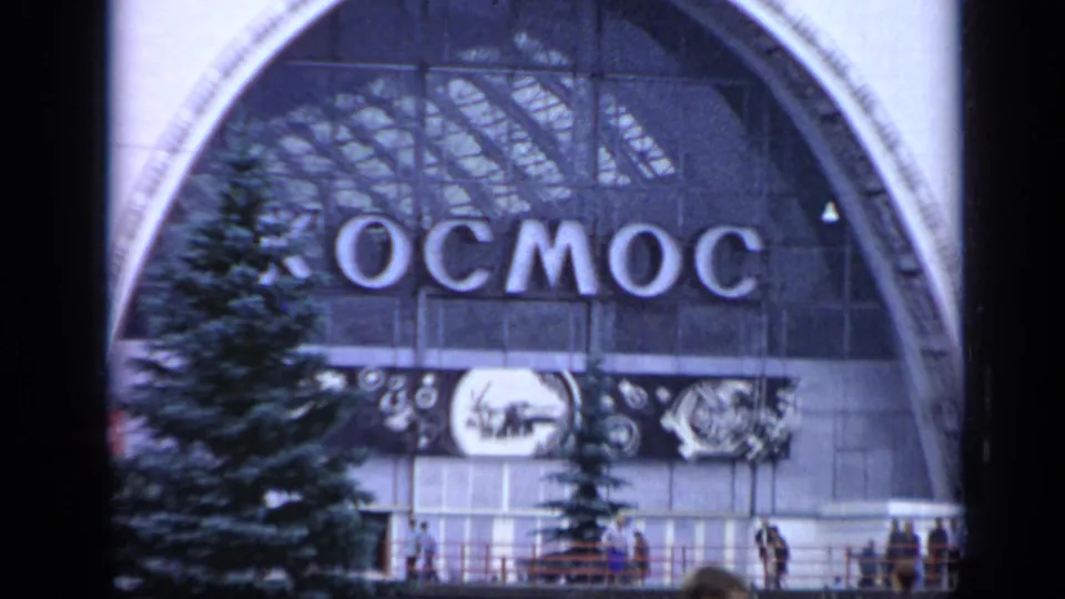 1971 Moscow home-movie frame of the Kosmos pavilion entrance, with arched glass, Cyrillic lettering, trees, and visitors in front.
