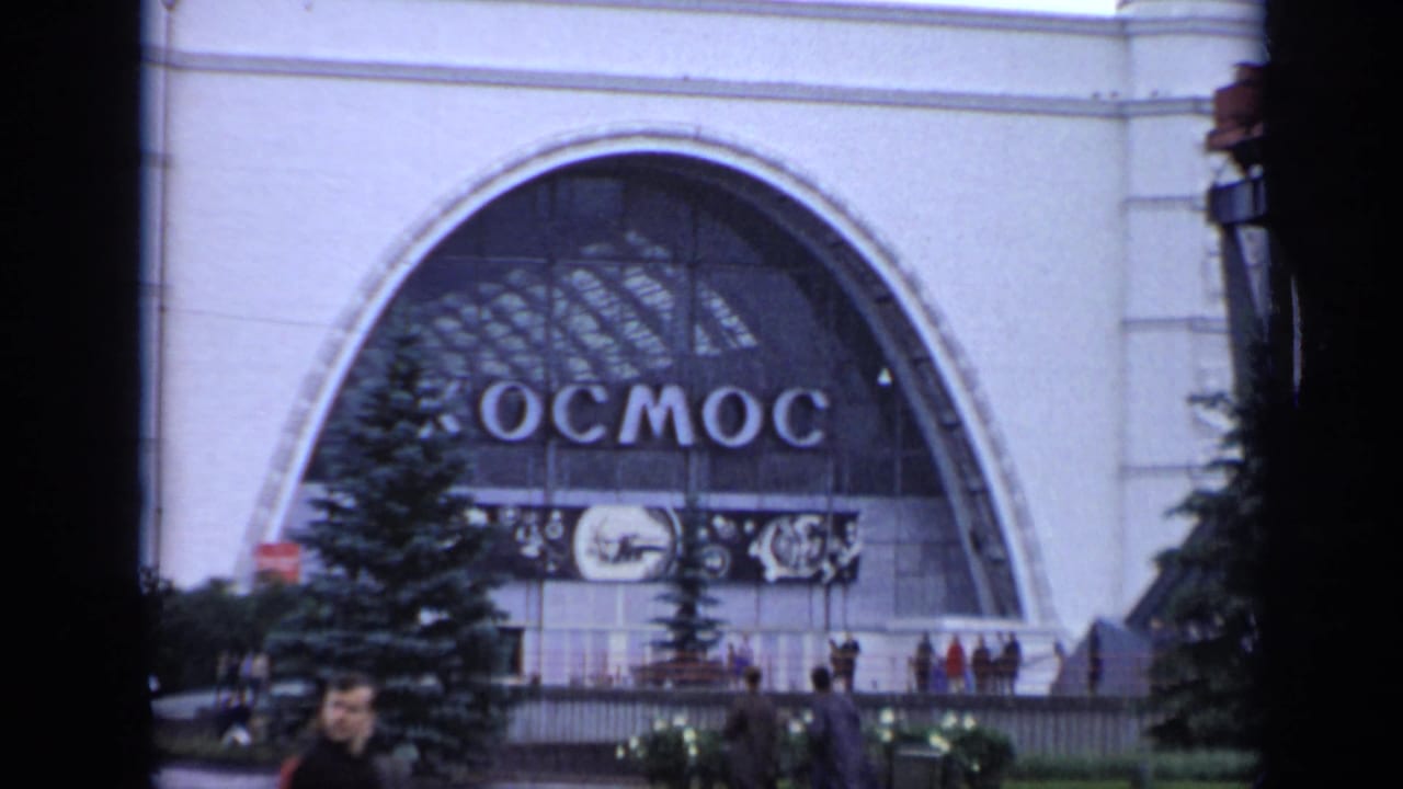 1971 Moscow frame showing the Kosmos pavilion from farther back, with visitors on the plaza and the large Cyrillic sign above the entrance.