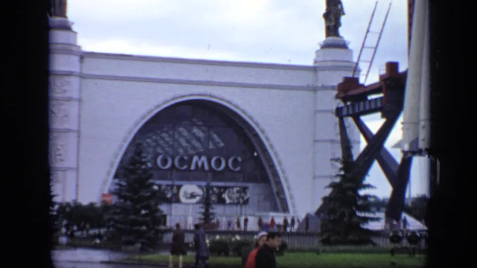 1971 Moscow frame of the Kosmos pavilion and nearby rocket display structure, with statues, cloudy sky, trees, and visitors in the foreground.