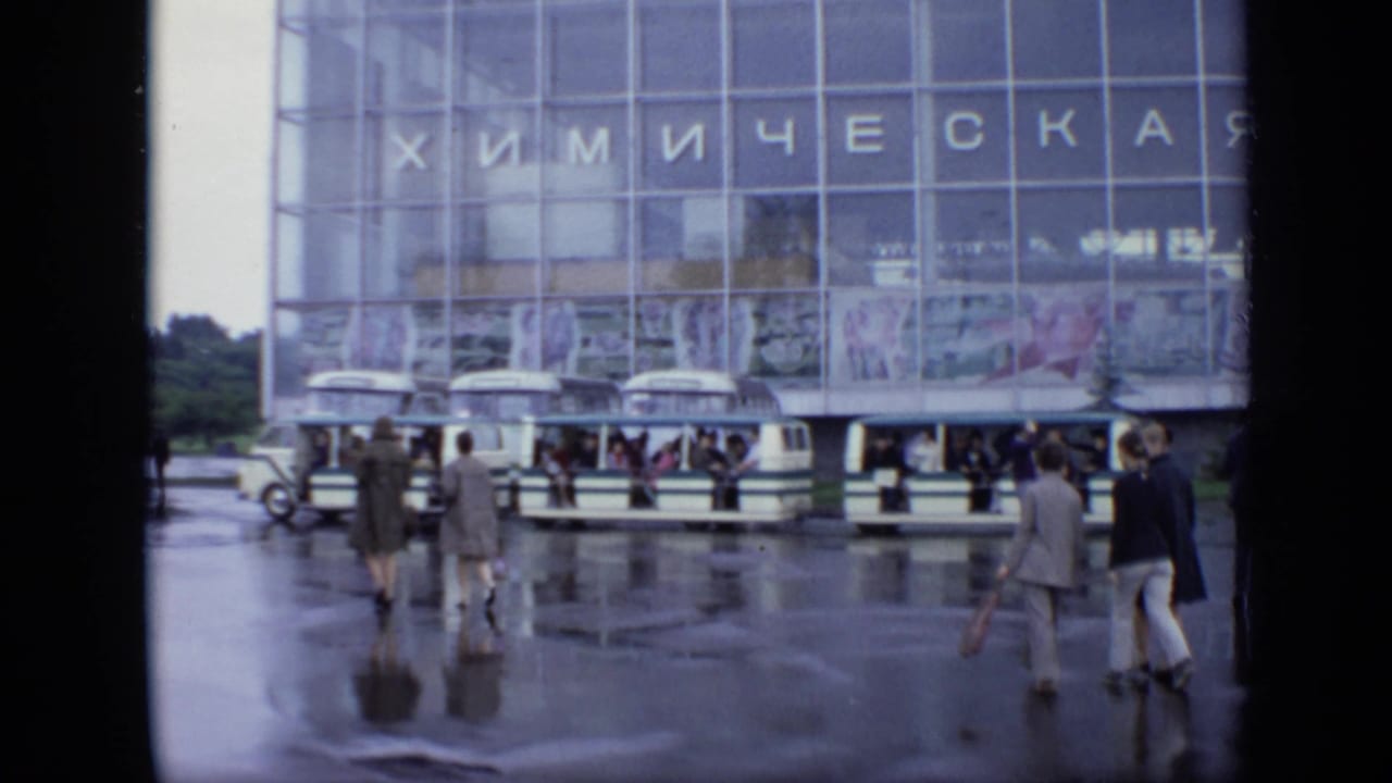 Pedestrians cross wet pavement in front of parked buses and a glass building with Cyrillic signage.