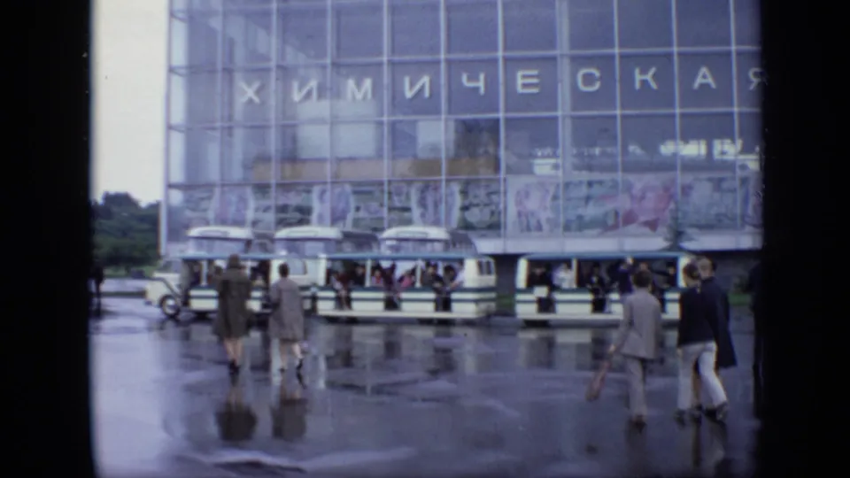 1971 Moscow pedestrians walk across rainy pavement near Soviet-era buses outside a glass-fronted building with Cyrillic text.
