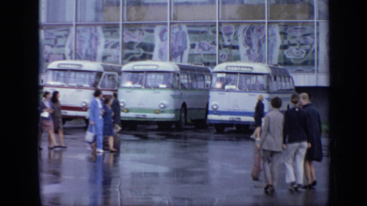 Several buses are parked outside a glass building while small groups of adults stand and walk nearby.