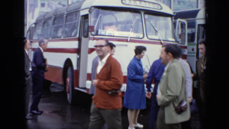 Adults move and wait beside a parked bus in rainy 1971 Moscow, captured in soft 8mm home-movie color.