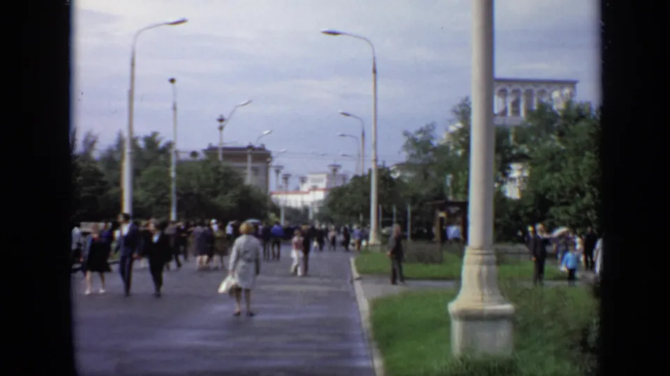 1971 Moscow street view with pedestrians on a wet tree-lined walkway, tall light poles, and distant buildings.