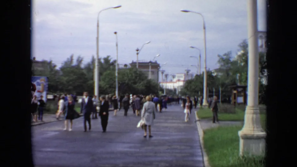1971 Moscow 8mm frame showing people spread across a wet pedestrian walkway bordered by trees and light poles.