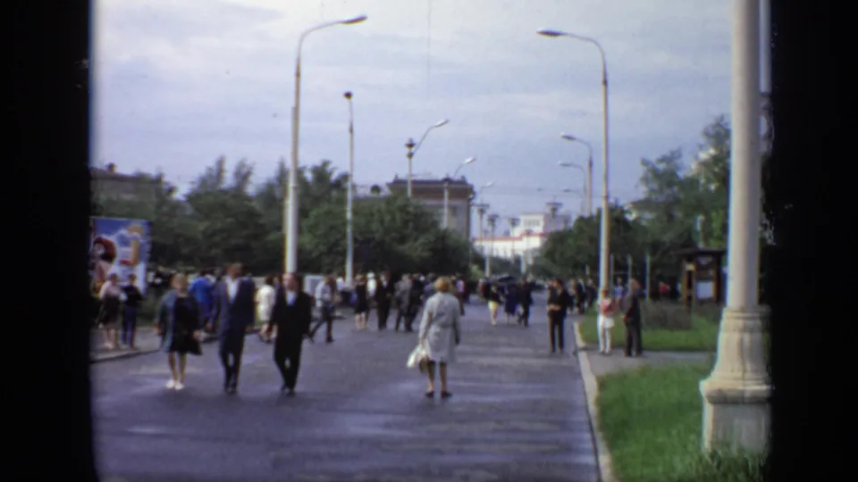 1971 Moscow street footage frame with adults walking along a wet public path under tall streetlights.