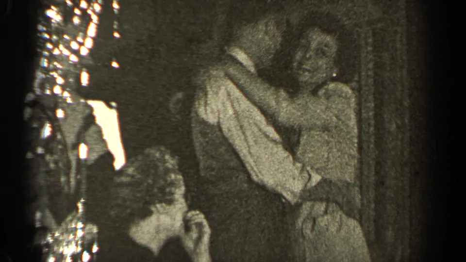 The couple embraces and kisses beneath a chandelier, with grain and motion blur reinforcing the 1947 home-movie feel.
